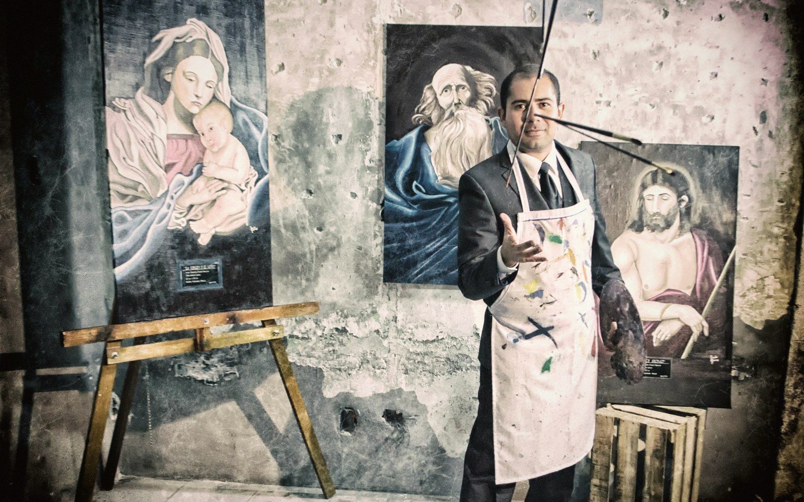 A man in a paint-splattered apron holds brushes in front of religious paintings on a weathered wall