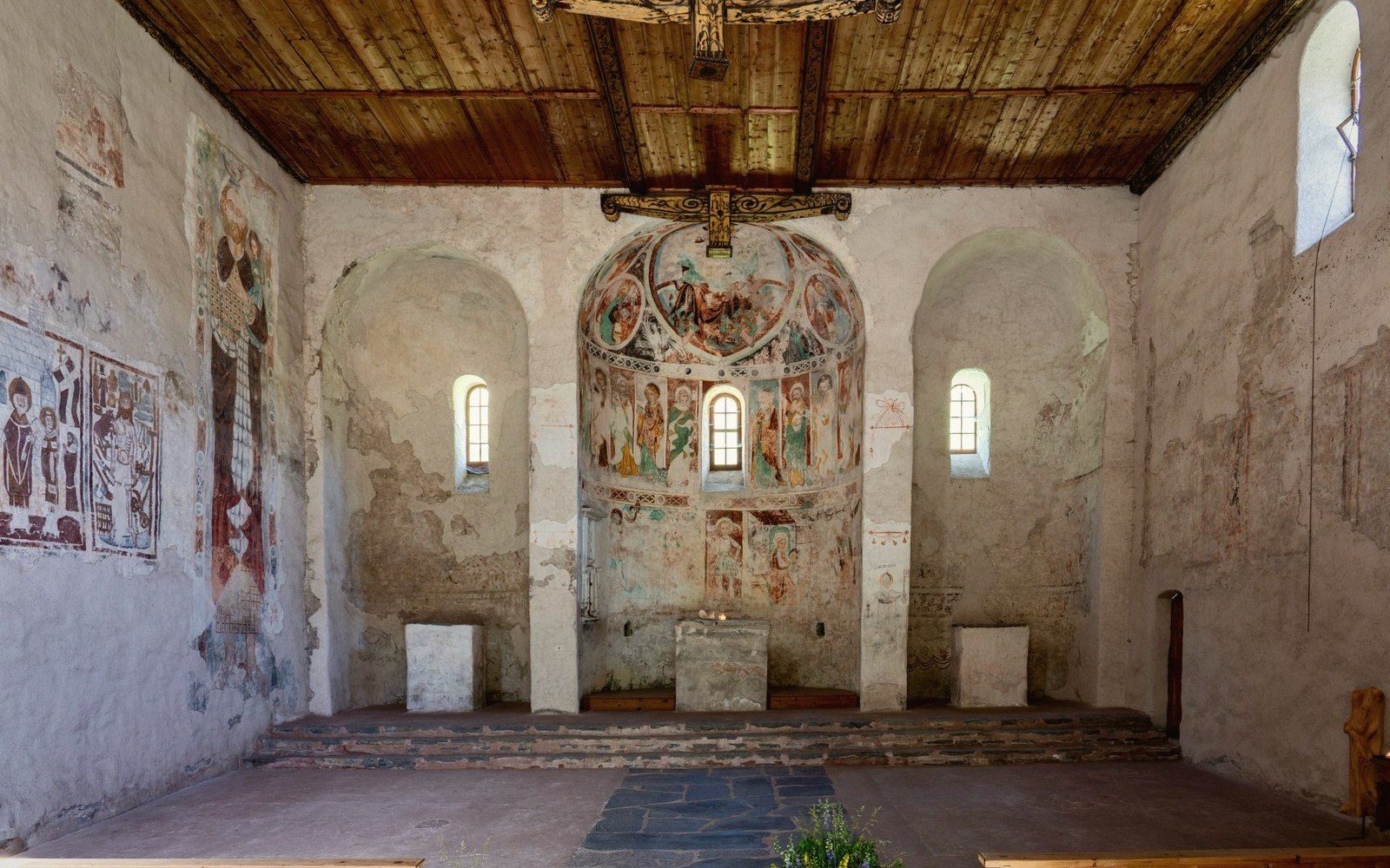 Faded medieval frescoes cover the apse walls of a small stone chapel with wooden ceiling